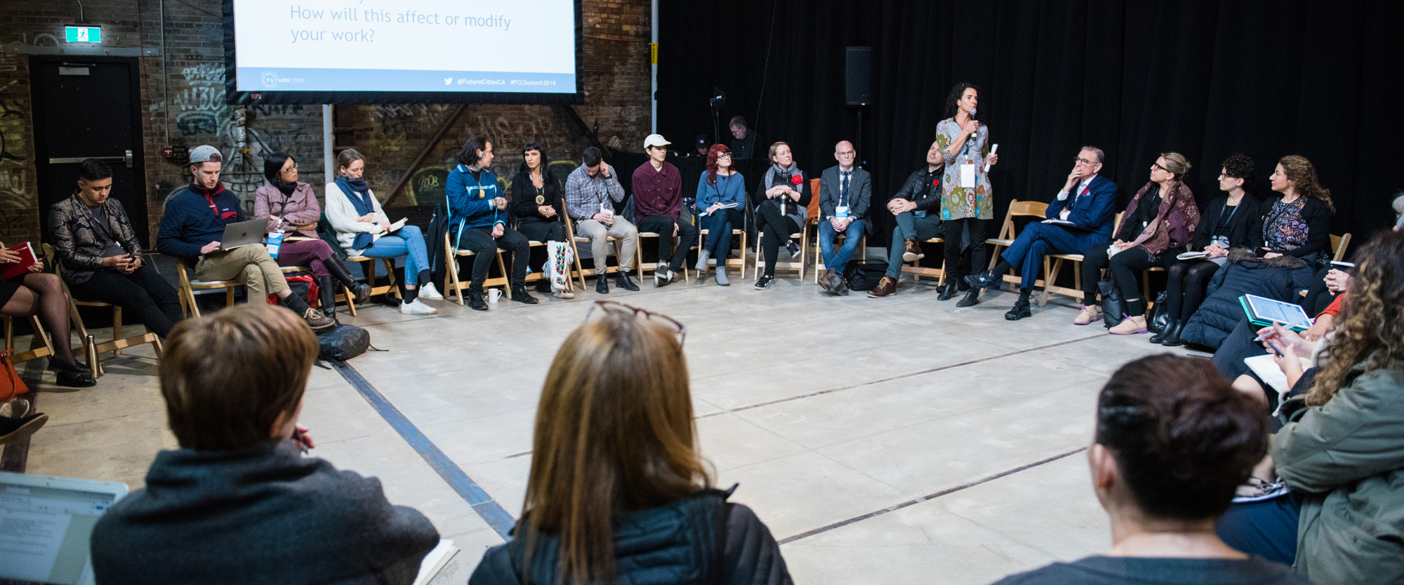 A group of people sit on on chairs in a circle during a discussion. One person stands speaking into a microphone. The group is on site at Evergreen Brick works in a building with brick walls.