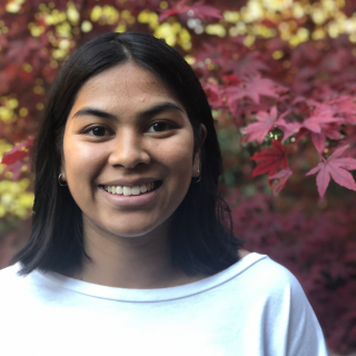 person with black hair wearing a white tshirt smiling broadly in front of fall foliage