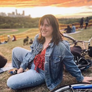 person wearing a bright red shirt with a jean jacket sitting next to a bike on a hill during sunset
