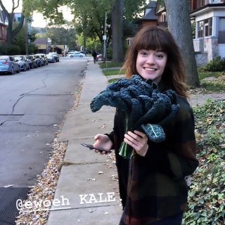 person wearing a plaid shirt holding a bunch of kale in one hand smiling at camera on a sidewalk