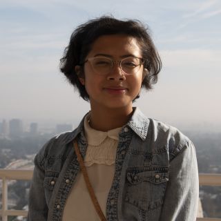 Person wearing glasses and a jean jacket looking into camera with a city skyline behind them