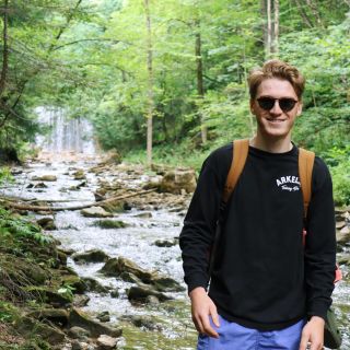 Person wearing sunglasses standing in a forest with a waterfall behind them