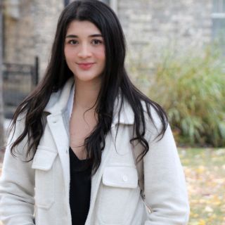 person with long dark hair wearing a white jacket standing in front of an old stone building