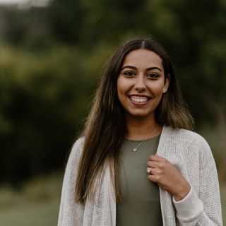 person with long hair wearing a green shirt and white cardigan smiling broadly in with trees behind them