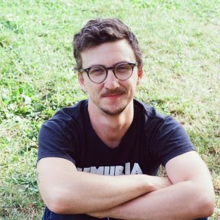 person wearing glasses and a tshirt with arms crossed smiling sitting on a field of grass