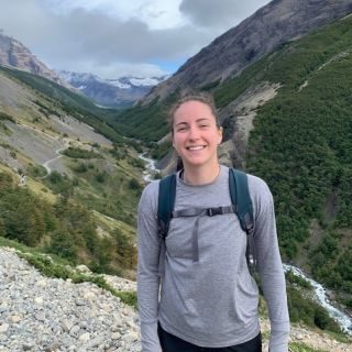 person wearing a grey shirt with a backpack standing and smiling in front of mountains