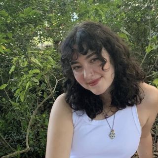 person wearing white tank top with brown curly hair smiling at camera in front of trees