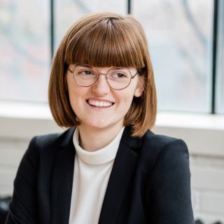 person wearing a cream shirt and black blazer with glasses and short ginger hair smiling