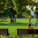 two people sitting on a park bench under the trees with family playing in the distance